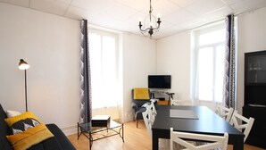 Een moderne woonkamer met een zwarte bank, een glazen salontafel, een eettafel met witte stoelen, een zwarte tv-kast en een kroonluchter.