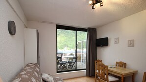 Et værelse med sofa, spisebord med stole, fjernsyn og et vindue med udsigt til grønne områder.