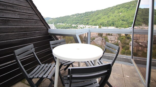 En balkon med et rundt bord og stole, med udsigt over en grøn bakke og en by.