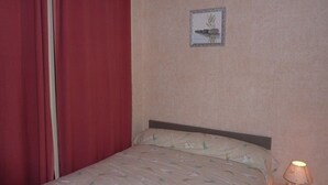 A hotel room with a bed, a nightstand with a lamp, a red curtain, and a framed picture on the wall. - Charming 1BR in Mont-Dore for 4, city center (Mont-Dore)