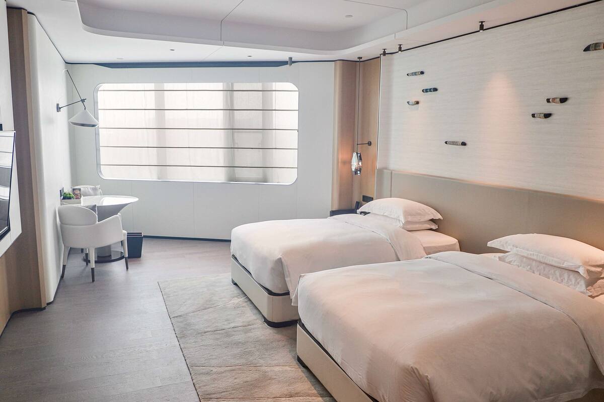 A modern hotel room with two beds, a desk, and a large window.