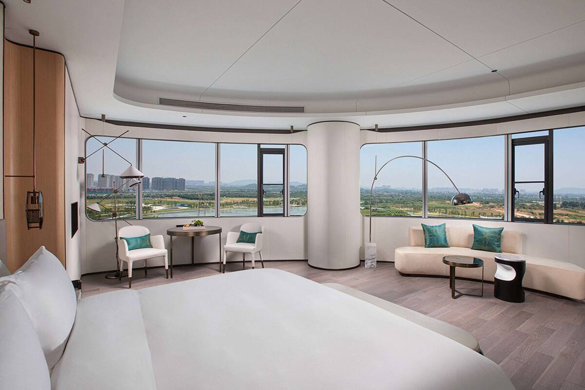 A modern hotel room with a large bed, a sofa, a dining area, and large windows offering a city view.