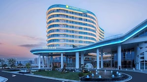 Un hotel moderno con fachada curva, una entrada destacada con toldo de cristal y un jardín ajardinado con fuente.