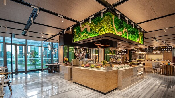 Un restaurante moderno con mostrador de recepción de madera, ventanas amplias y una pared decorativa verde.