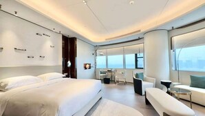 Una habitación de hotel moderna con una cama grande, una zona de estar con un sofá y sillones, una mesita y un ventanal con vistas a la ciudad.