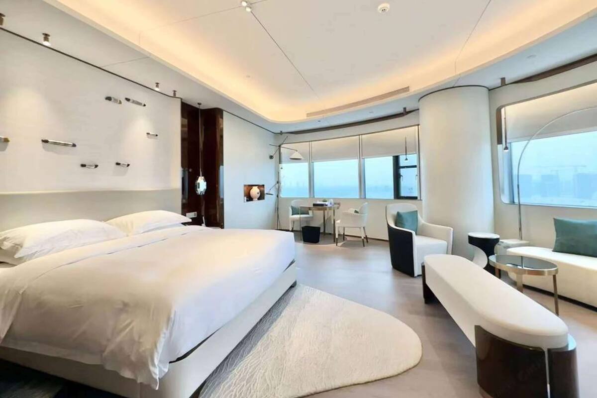 A modern hotel room with a large bed, a seating area with a sofa and chairs, a small table, and a large window offering a city view.