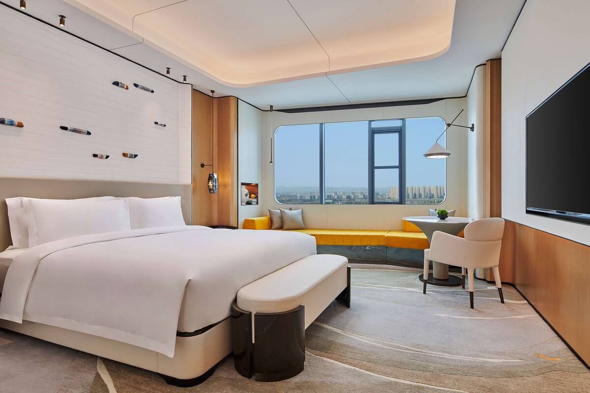 A modern hotel room with a large bed, a small desk, and a view of the city through large windows.