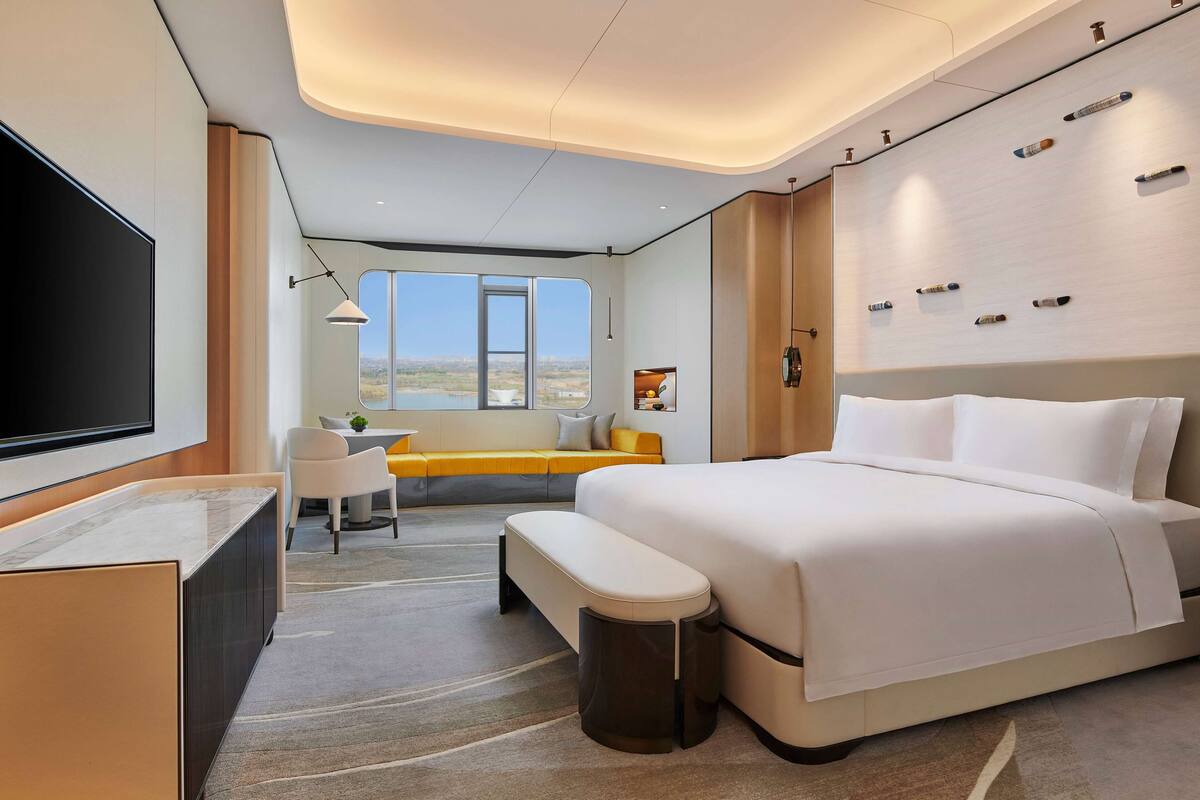 A modern hotel room with a large bed, a desk, a seating area with a view, and a TV mounted on the wall.
