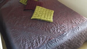 Ein Bett mit dunkler Bettdecke und drei Kissen in verschiedenen Farben. Ein Nachttisch mit Lampe und kleiner Schublade steht daneben. Über dem Bett hängt ein Bild.