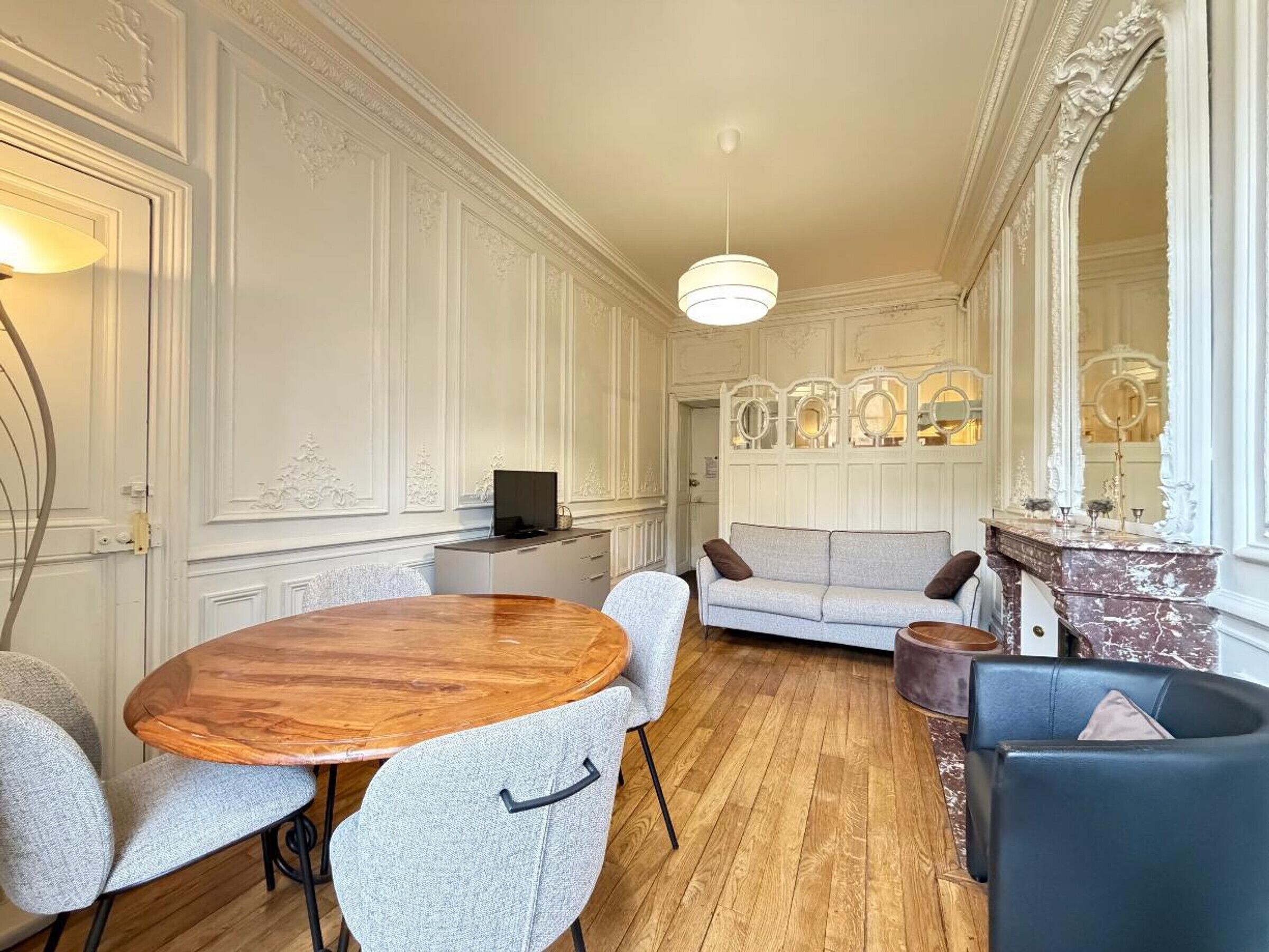 Um quarto com uma mesa de madeira redonda, cadeiras estofadas cinzentas, um sofá, uma lareira e um espelho grande.