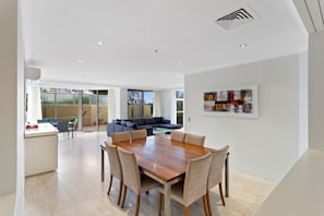 A spacious dining area with a wooden table and chairs, a living room with a sofa and a dining table, and a large window providing a view of the outdoors.