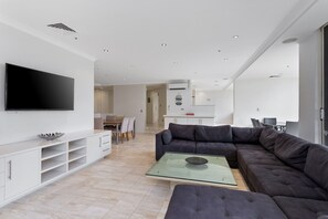 A modern living room with a flat-screen TV, a glass coffee table, a dark sofa, and a dining area with a table and chairs. - Spacious 3 Bedroom Family Apartment (Surfers Paradise)