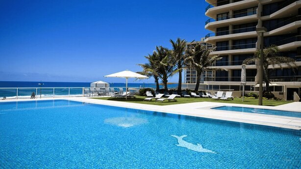A clear blue pool with lounge chairs and umbrellas, a palm tree-lined deck, and a view of the ocean.