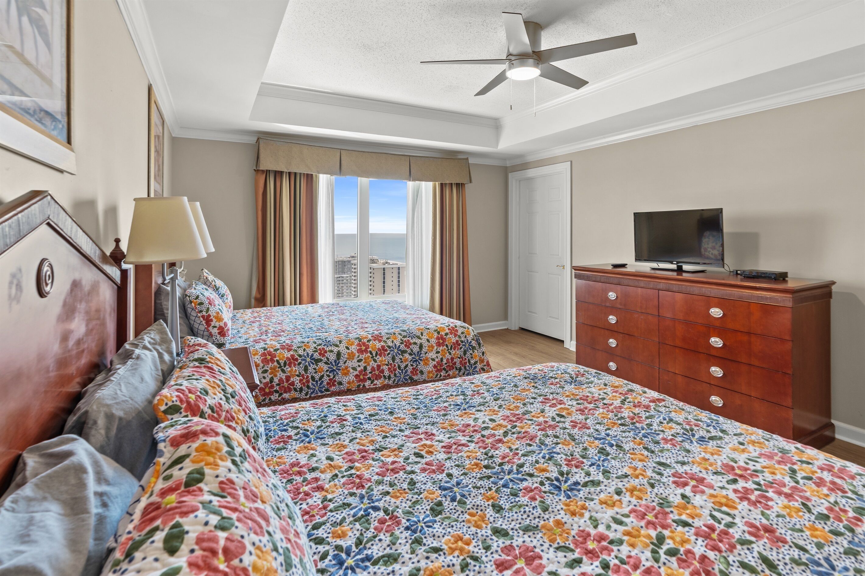 Quarto de hotel com duas camas, televisão, penteadeira e uma janela grande com cortinas.