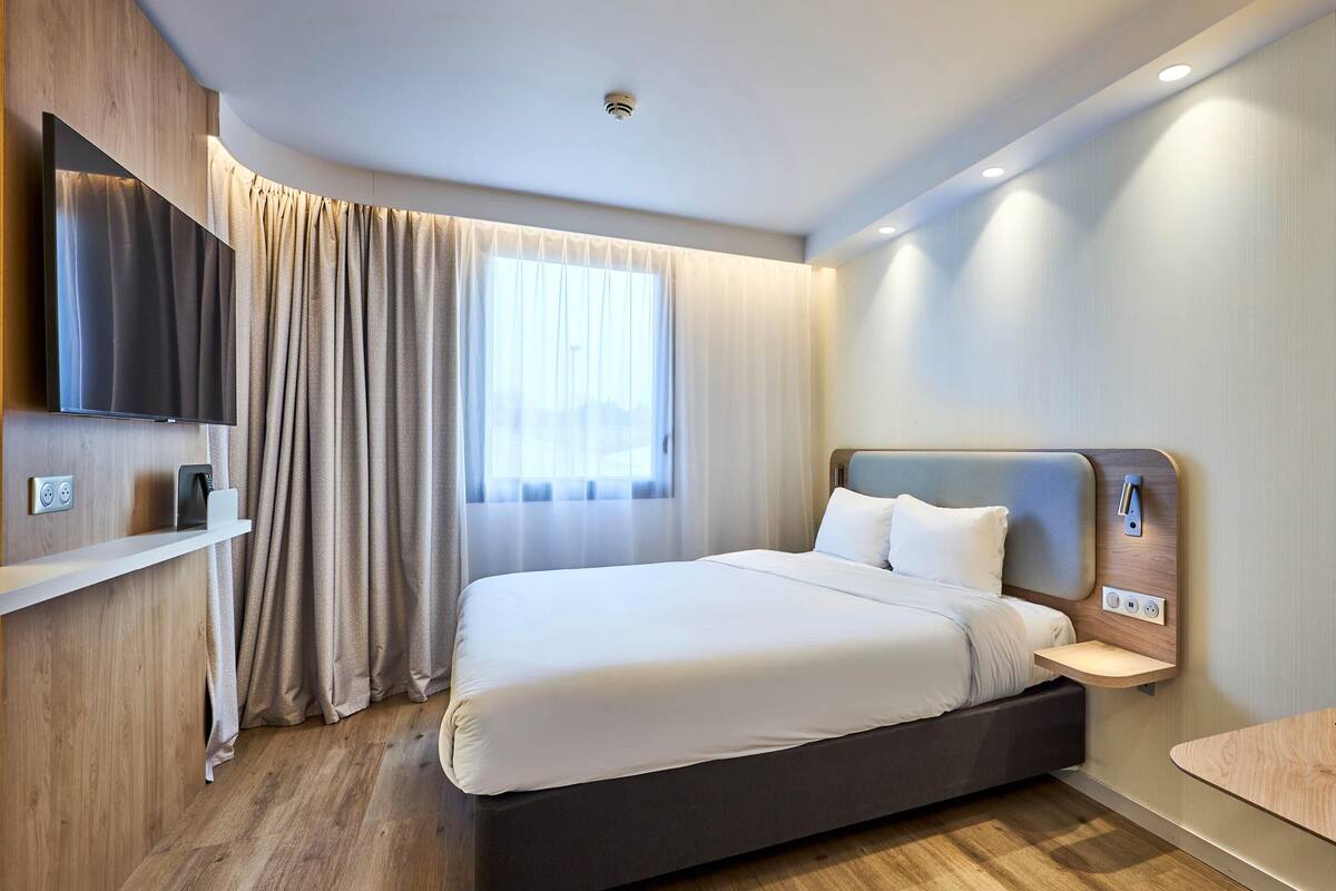 A modern hotel room with a large bed, a mounted TV, a bedside table, and a window with curtains.
