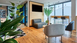 A modern hotel lobby with a wooden floor, a grey sofa, a television mounted on a wooden wall, and a large window offering a view of the outside. - B&B HOTEL Deauville-Touques (Touques)