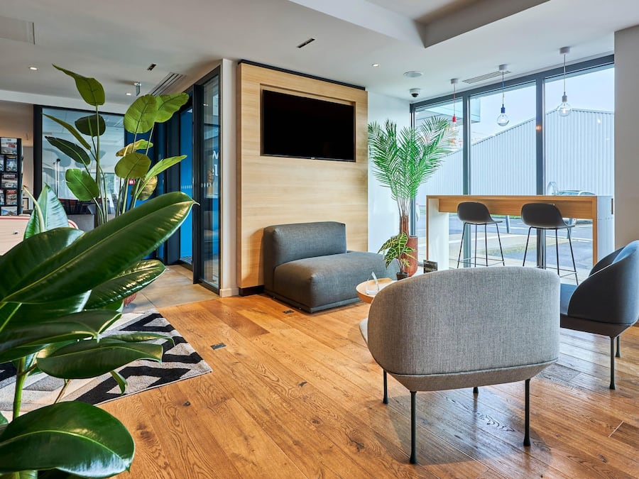 Eine moderne Hotellobby mit Holzboden, einem grauen Sofa, einem an einer Holzwand montierten Fernseher und einem großen Fenster mit Blick ins Freie.