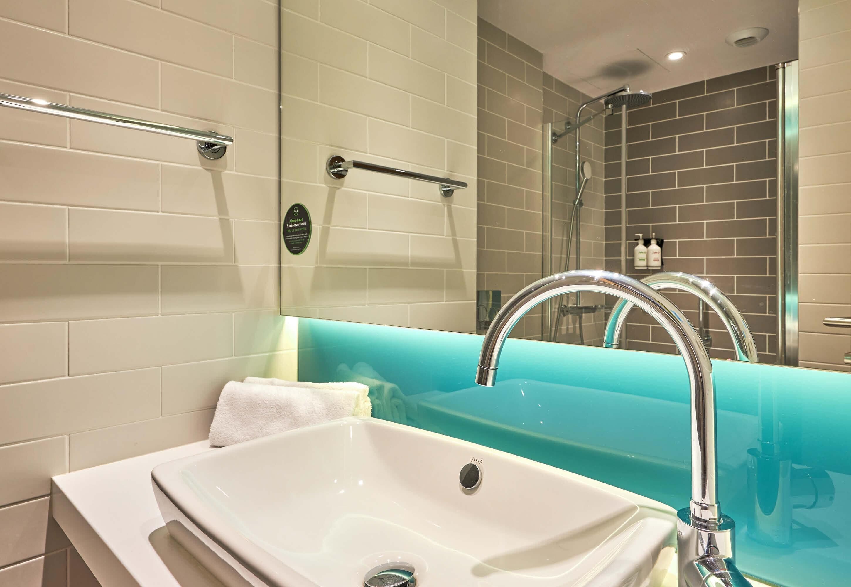 Un baño con un lavabo blanco, un grifo cromado, un perchero y un espejo que refleja la zona de la ducha.