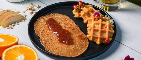 Un plato con un panqueque, un waffle y fruta cortada sobre una mesa de madera blanca.