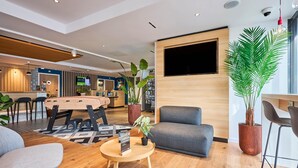 A modern hotel lobby with a wooden reception desk, comfortable seating, a large TV screen, and potted plants. - B&B HOTEL Deauville-Touques (Touques)