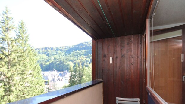 Un balcón con una mesa y sillas rojas, paredes de madera y vistas a un bosque y edificios.