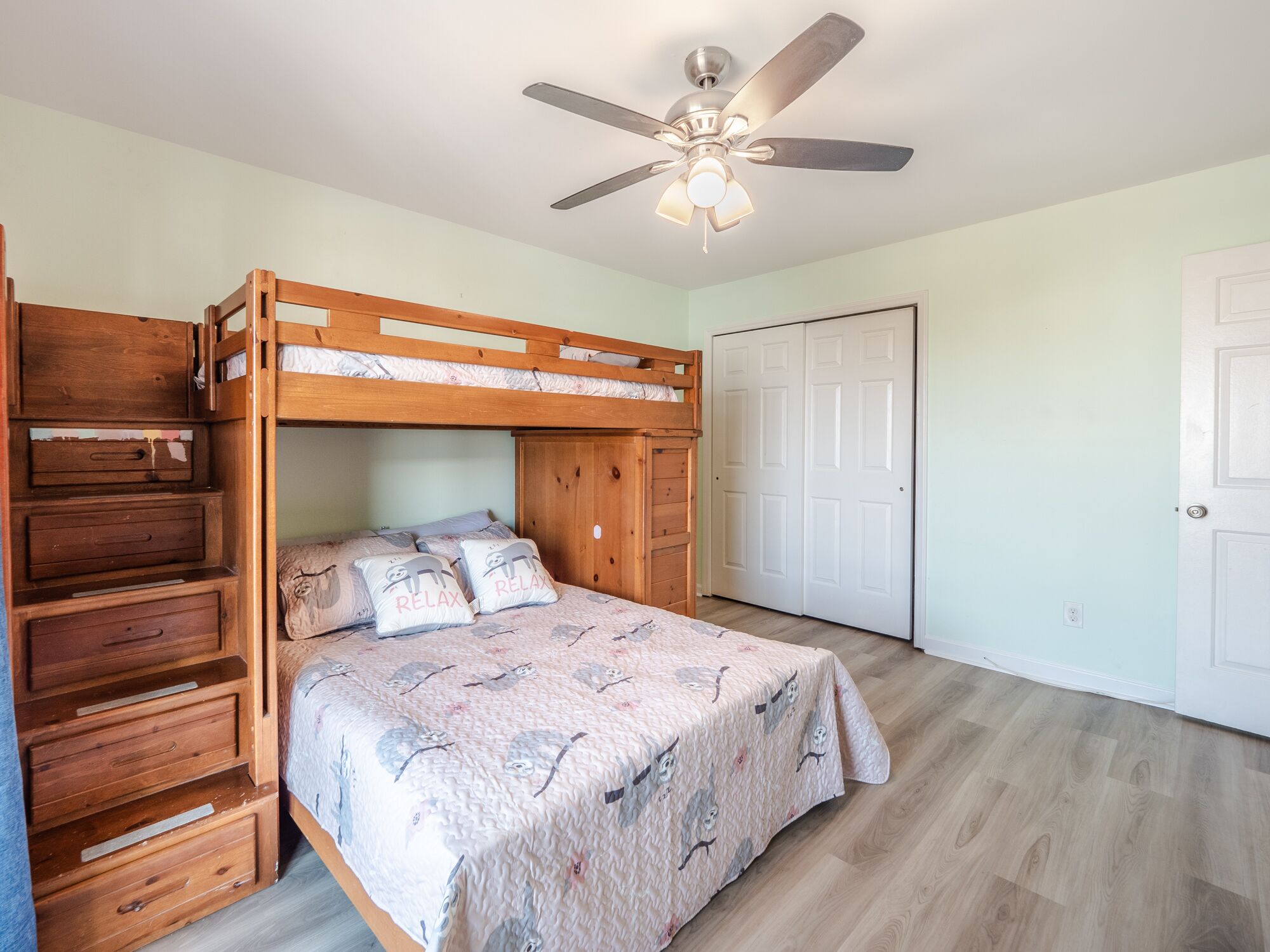 Ein Stockbett mit Holzlade und Schubladen, ein Deckenventilator und eine Tür in einem Zimmer mit hellen Wänden und Holzboden.