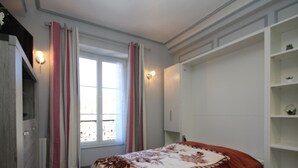 Et soveværelse med seng, et vindue med gardiner, et skab og en reol med planter.