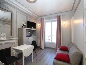 Quarto compacto com uma mesa branca, uma cadeira preta, um sofá cinza com almofadas vermelhas, um armário de madeira, uma TV em cima e uma janela com cortinas.