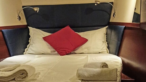 Ein ordentlich bezogenes Bett mit dunkelbraunem Kopfteil, weißen und roten Kissen sowie einer beigen Decke.