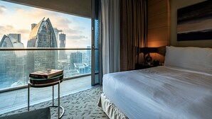 Une chambre d’hôtel avec un grand lit, une petite table sur laquelle se trouve un livre, une table de chevet avec une lampe, et une vue sur le paysage urbain par une grande fenêtre.