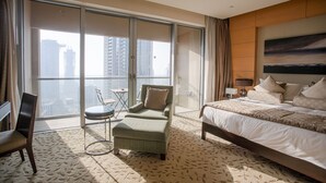 Une chambre d’hôtel dotée d’un grand lit, d’un coin salon avec un fauteuil et un pouf, d’une petite table et offrant une vue sur la ville grâce à de grandes fenêtres.