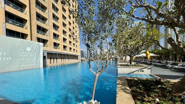 Une piscine avec un arbre et des fleurs au premier plan, entourée de chaises longues et d’un bâtiment sur lequel est accrochée une banderole.