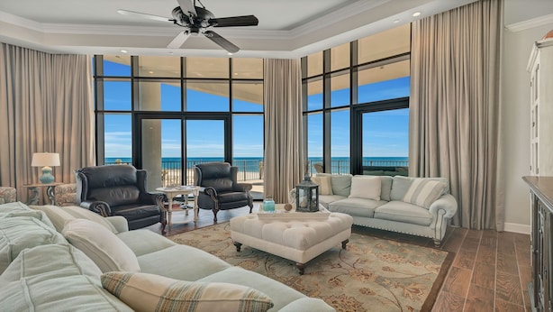 A spacious living room with a large window overlooking the beach, leather armchairs, a sofa, and a ceiling fan.