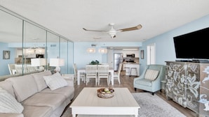A modern living room with a sofa, coffee table, and a TV. There's a dining area with chairs and a table, and a kitchen in the background. - New to the Rental Market, New Owner, Newly Renovated!!! (Orange Beach)
