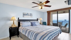 Une chambre à coucher avec un lit, deux tables de chevet avec des lampes, un ventilateur de plafond, un tableau au mur et un balcon avec vue sur l’océan.