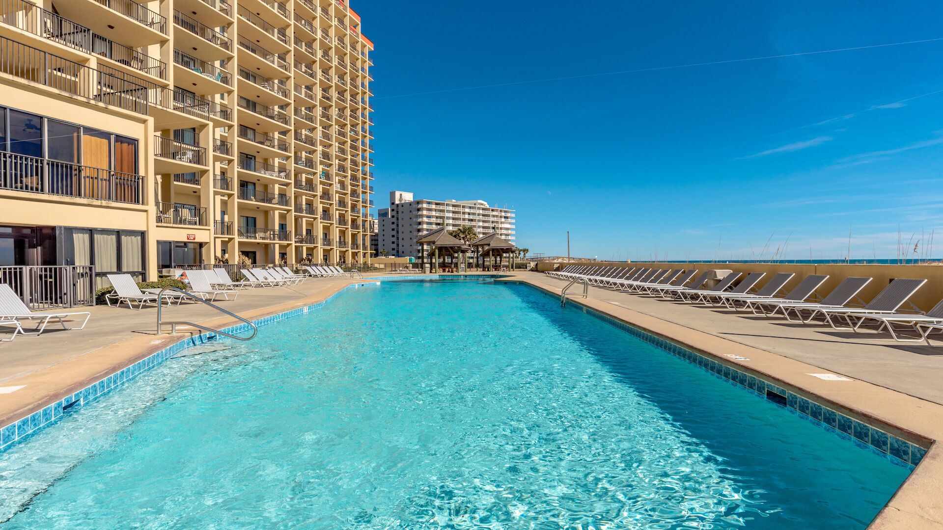 Piscina com espreguiçadeiras em ambos os lados, um edifício com vários andares ao fundo e céu claro.