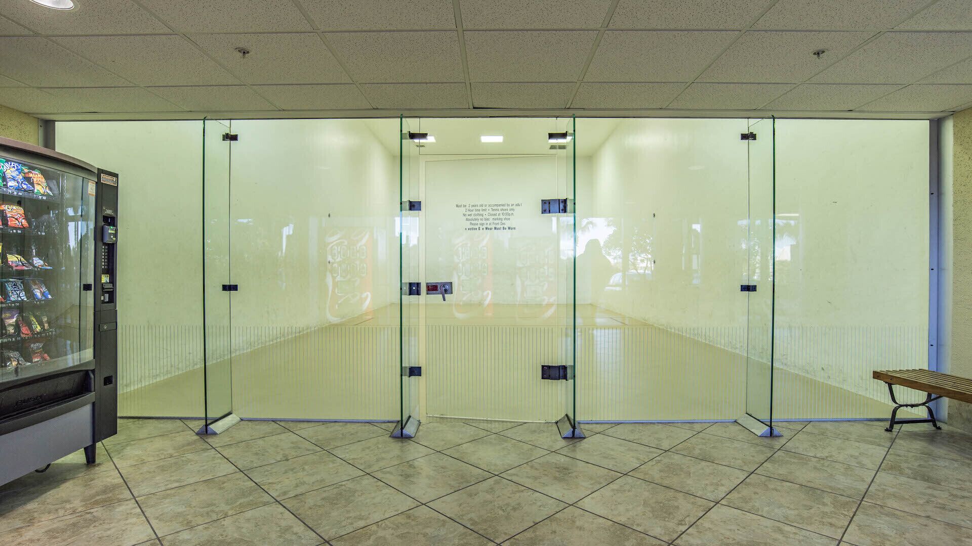 Un court de squash avec des murs en verre, un distributeur automatique et un banc.