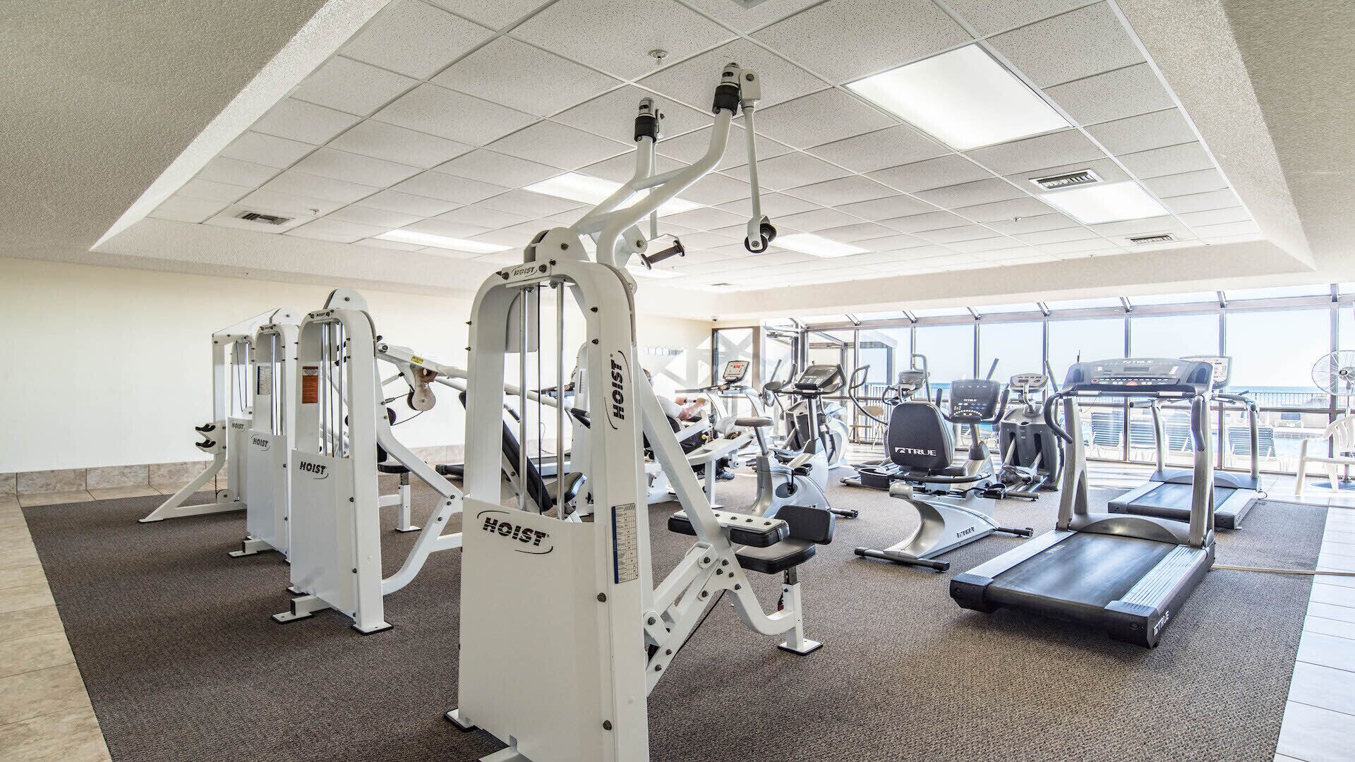 Une salle de sport équipée de divers appareils d’exercice, notamment des machines de musculation et des tapis de course, et dotée de grandes fenêtres offrant une vue dégagée.