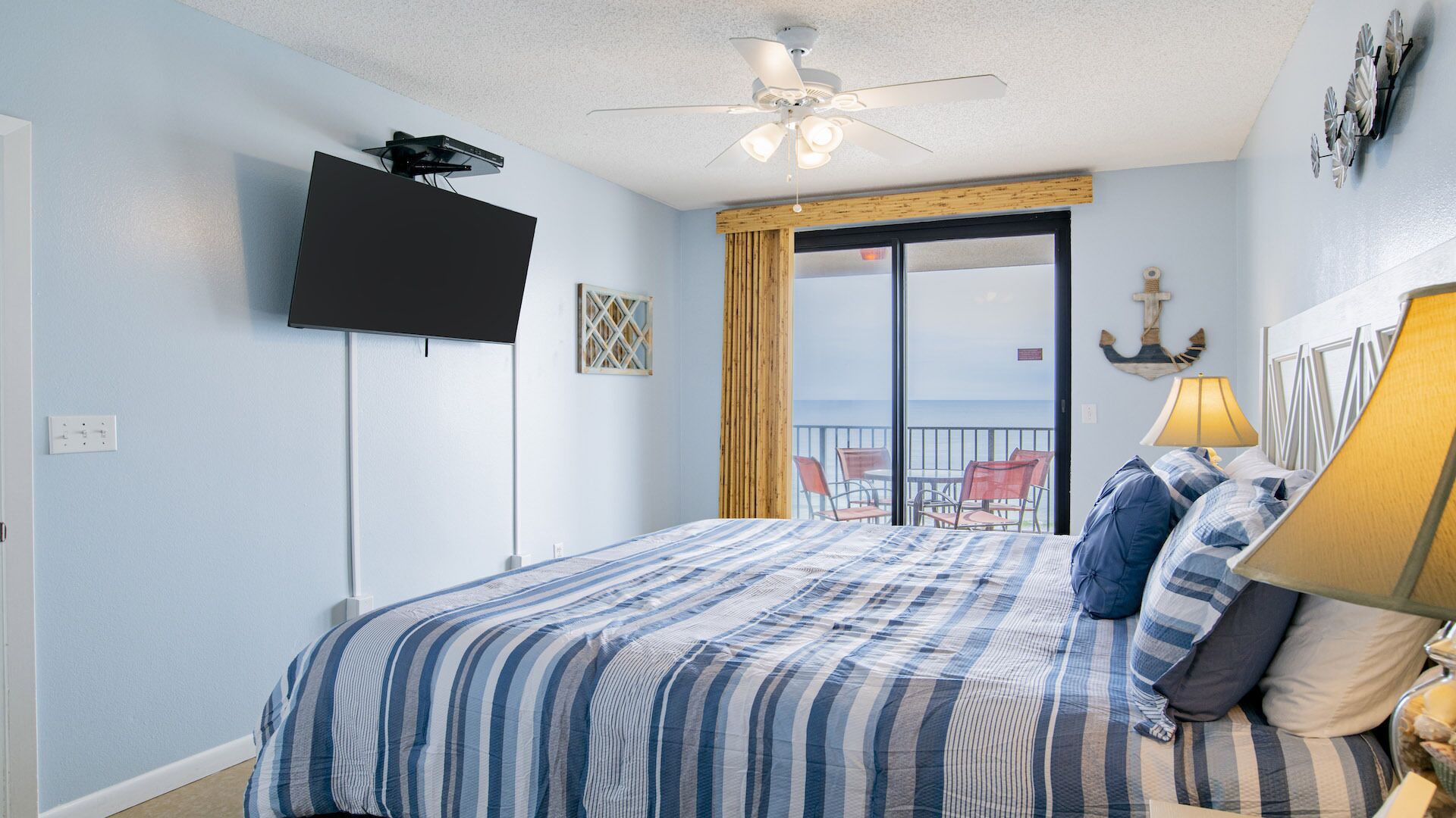 A bedroom with a bed, a television mounted on the wall, a ceiling fan, and a view of the ocean through a sliding glass door.