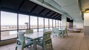 A spacious indoor area with large windows offering a view of the beach and cityscape. There are several wooden tables and chairs arranged for dining or socializing.
