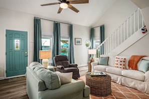 Uma sala de estar com uma porta verde-água, dois sofás, uma poltrona escura, um ventilador de teto e uma escada.