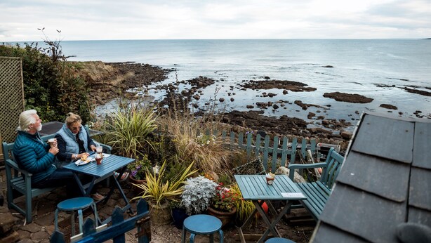 Two people seated at a table with a view of the sea and rocky shoreline.
