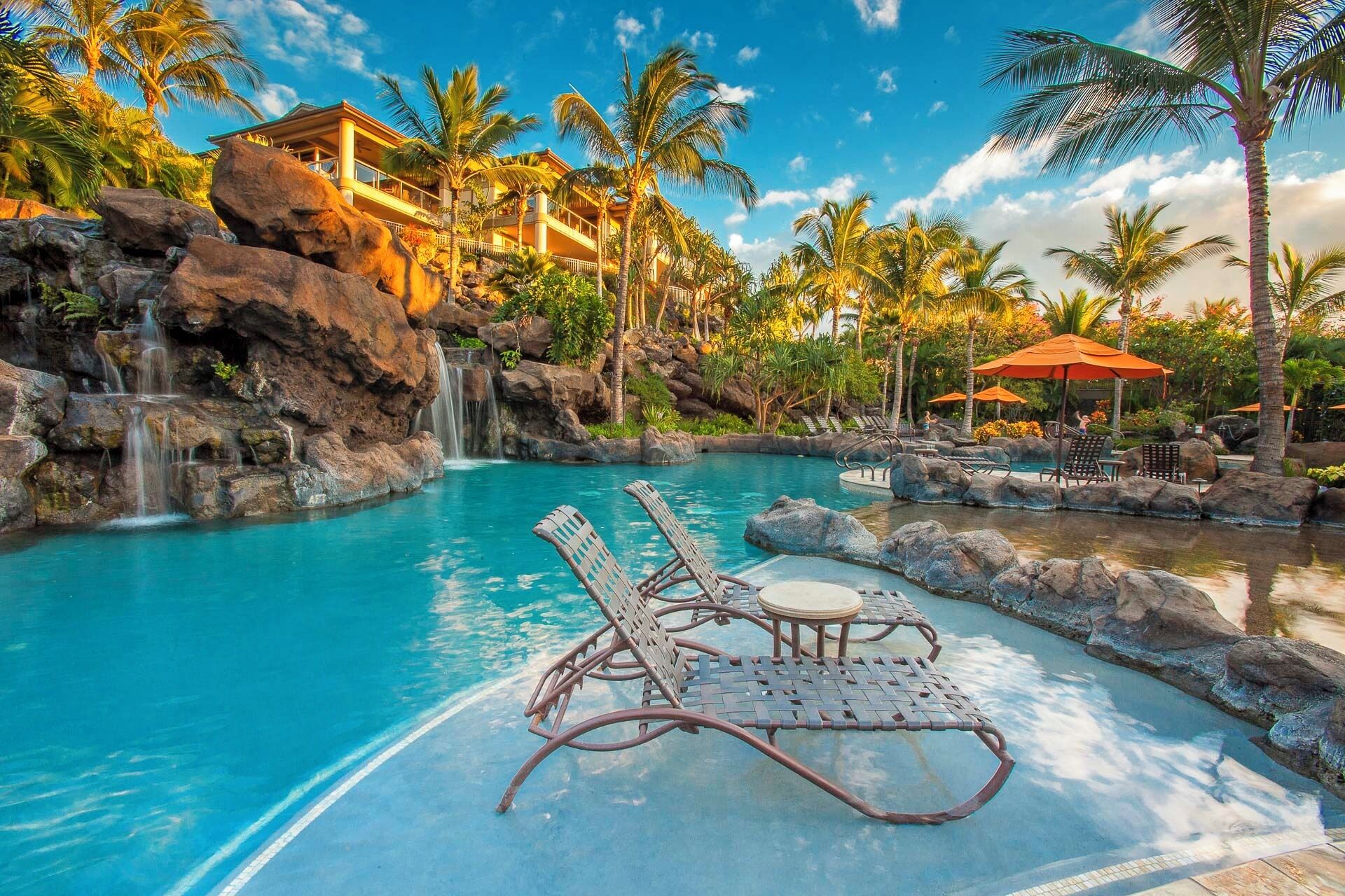 Piscina de un complejo turístico con sillas de descanso y sombrillas, rodeada de palmeras y una cascada.