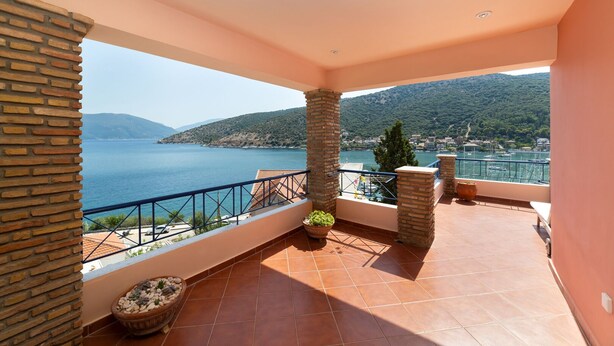 A balcony with a view of a lake and mountains, featuring a tiled floor, a railing, and potted plants.