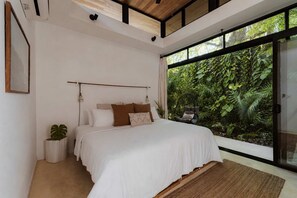 A bedroom with a large bed, a hanging chair, a potted plant, and a view of lush greenery through large windows.
