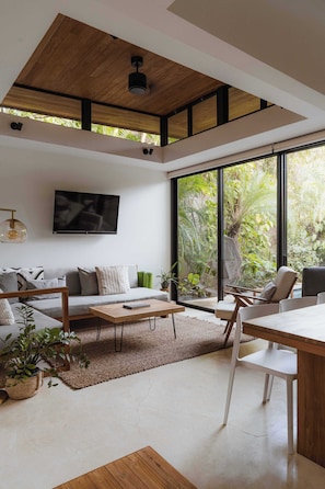 Una sala de estar moderna con un sofá, una mesa de centro y sillas, ventanas amplias con vistas a zonas verdes y un televisor de pantalla plana montado en la pared.