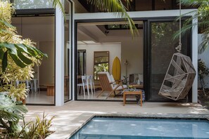 Ett modernt hus med en glasdörr som leder ut till ett poolområde, där det finns en stol, en surfbräda och en hängstol.