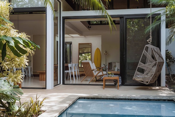 Una casa moderna con puerta de vidrio que da a un área de piscina, con una silla, una tabla de surf y un sillón colgante.