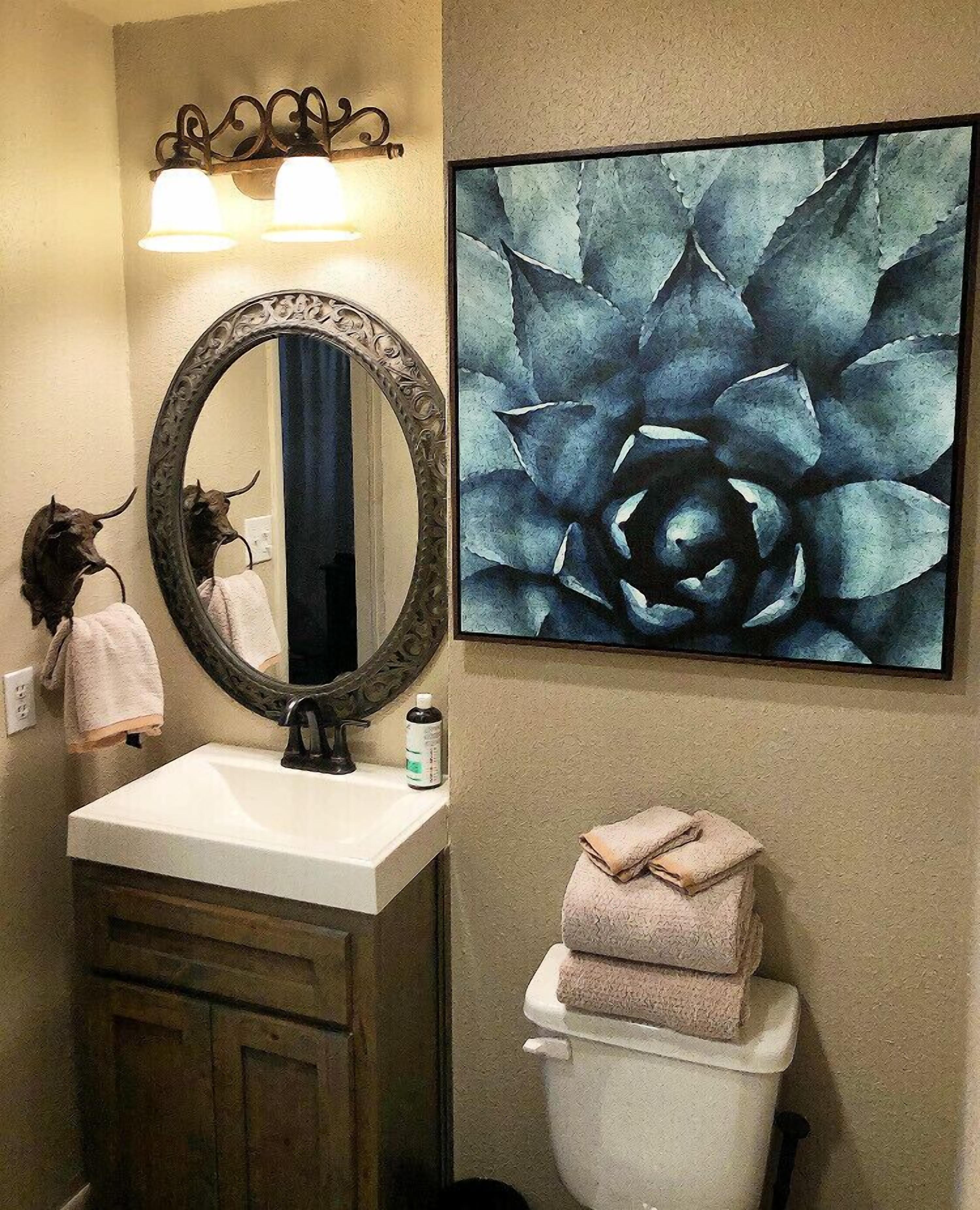 A bathroom with a white sink, a large ornate mirror, a wall-mounted light fixture, a framed abstract painting, and a toilet with stacked towels.
