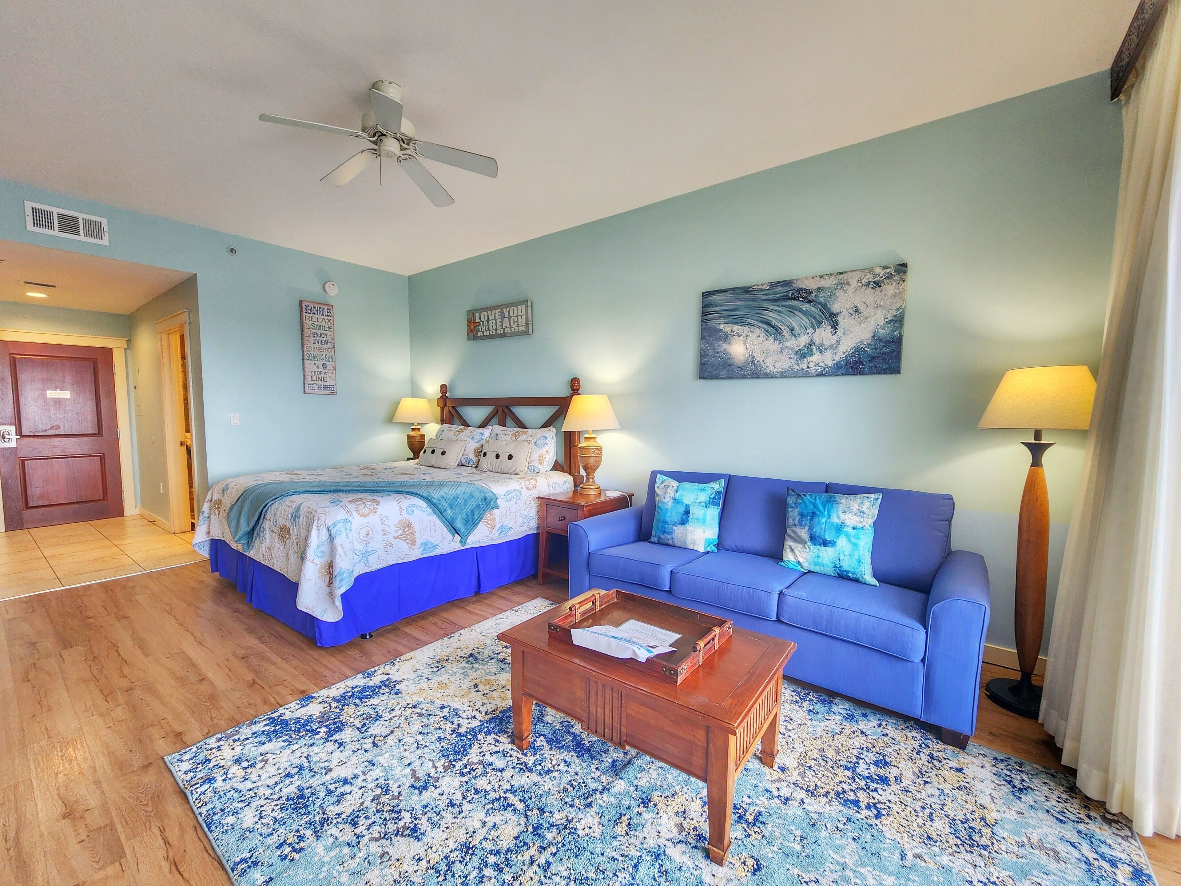Een slaapkamer met een bed, een blauwe bank, een houten salontafel en een plafondventilator.